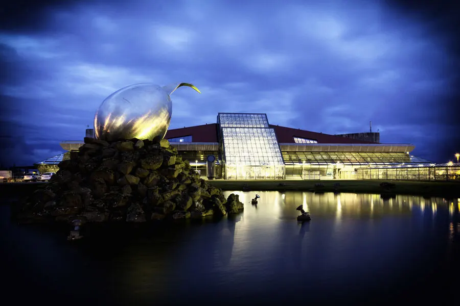 Keflavík International Airport terminal building lit up at dusk with its distinctive stainless steel sculpture in front