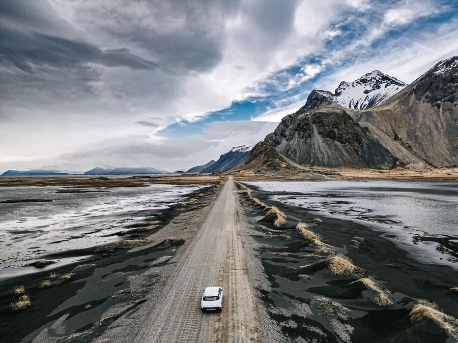 Aerial view of a single car driving a long gravel road across Iceland's black sand plains toward snow-capped mountains