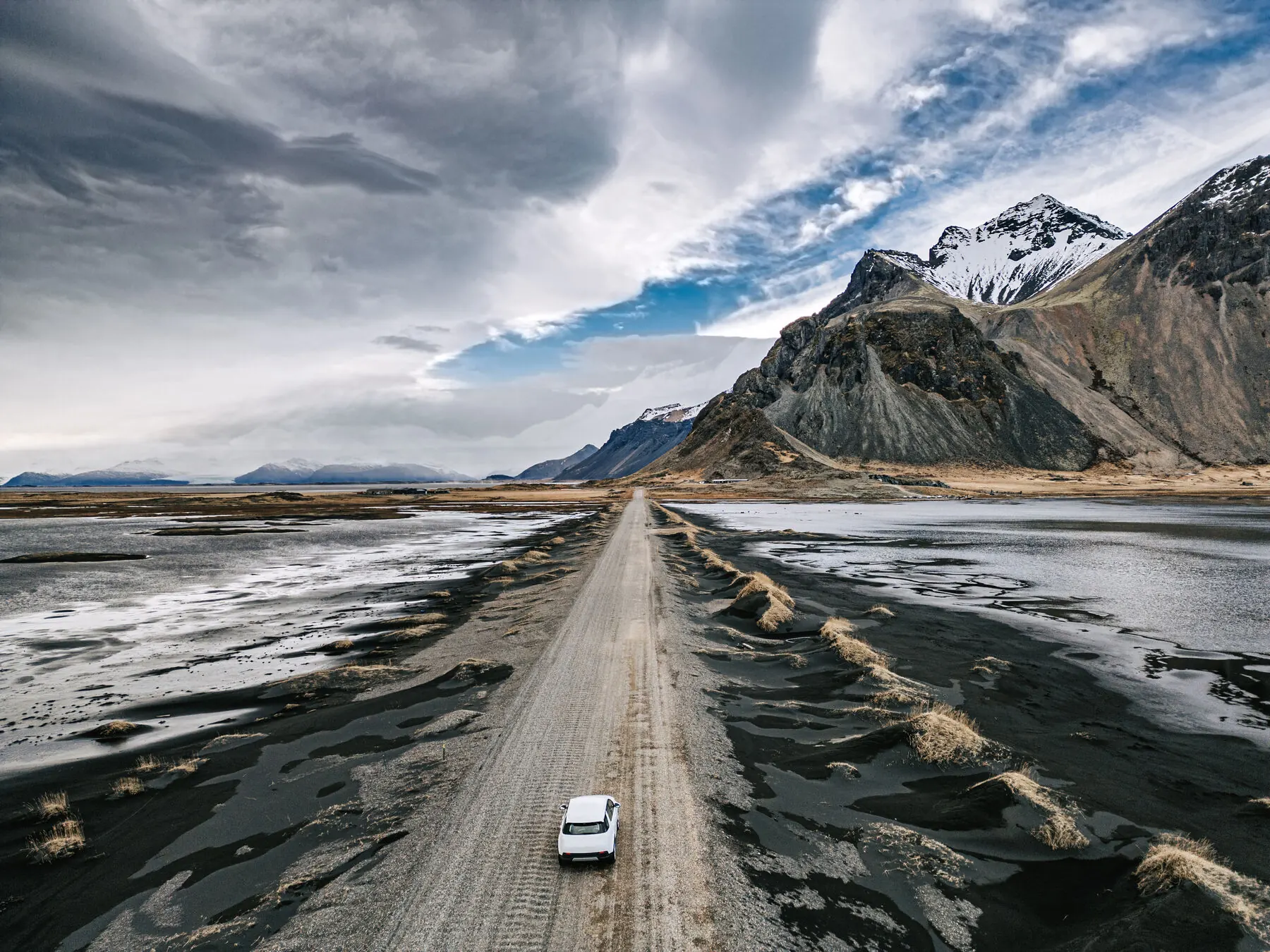 Aerial view of a single car driving a long gravel road across Iceland's black sand plains toward snow-capped mountains