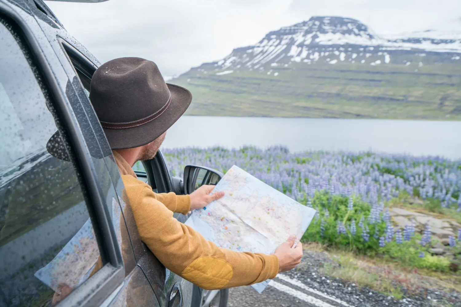A traveler leaning out of a car window studying a paper map of Iceland beside a lake and mountains