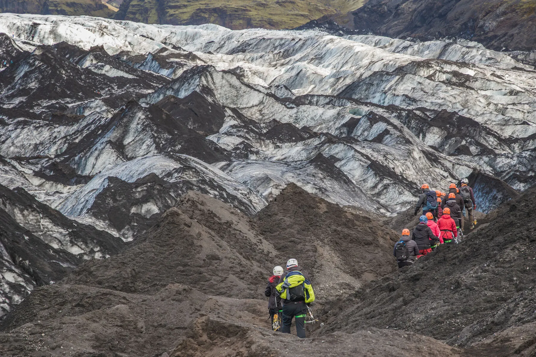 Une cordée de randonneurs casqués et équipés de crampons traversant un glacier crevassé en Islande, avec un groupe descendant une crête de cendres volcaniques au premier plan