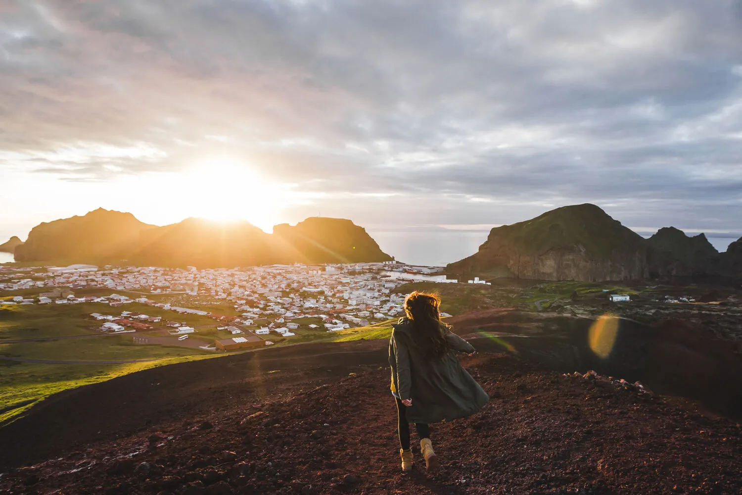 Un randonneur marchant sur la crête volcanique de l’Eldfell au coucher du soleil, avec la ville de Vestmannaeyjar en contrebas