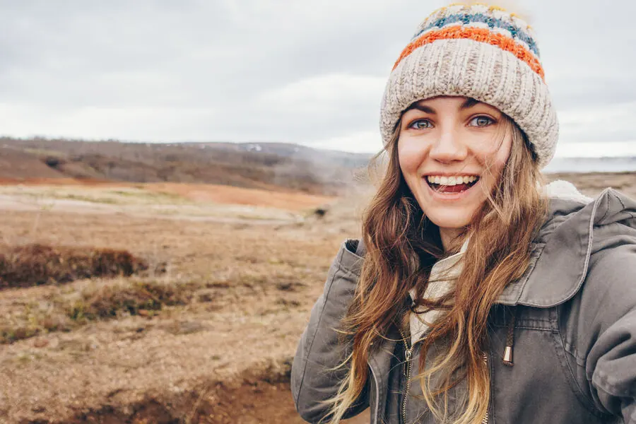 A traveler bundled in a warm hat and jacket, smiling in a windswept Icelandic geothermal landscape